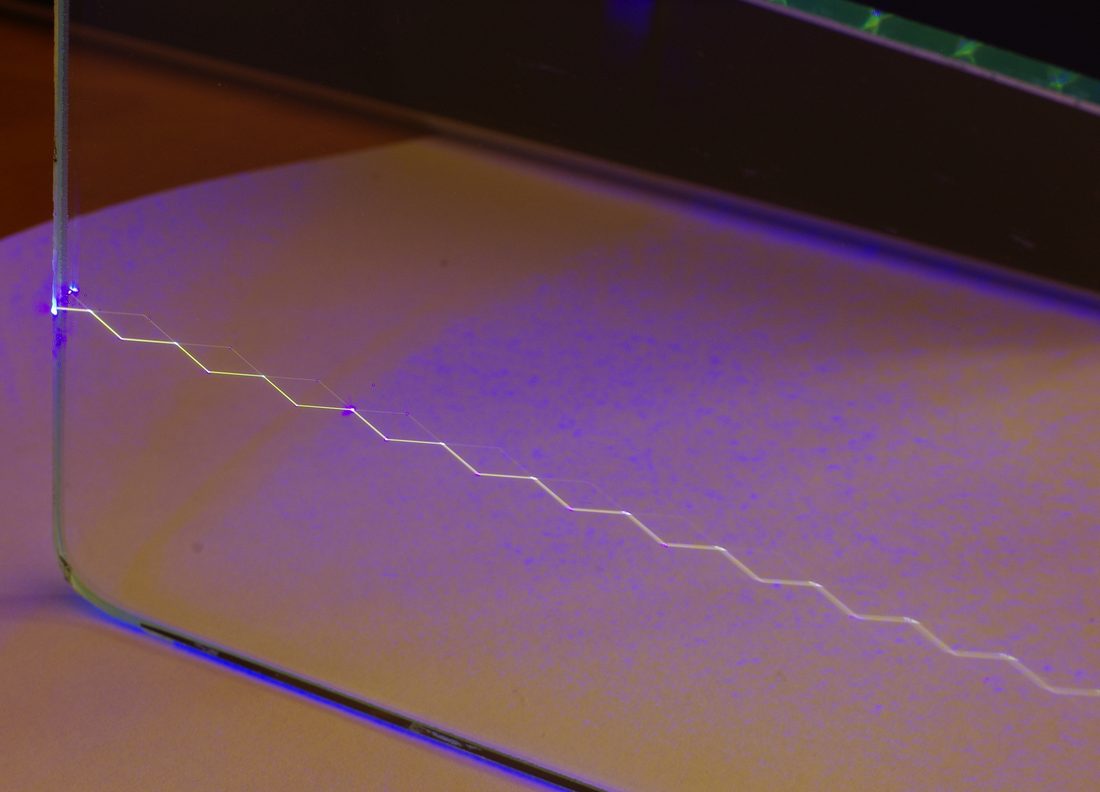 Kalın bir cam levhanın kenarından giren 405 nm’lik mor lazer, tam yansımayla zikzak çizerek ilerliyor ve geçtiği yeri yeşilimsi parlatıyor. Işığı içeride tutan şeyin ceket değil geliş açısı olduğunu doğrudan gösteriyor.